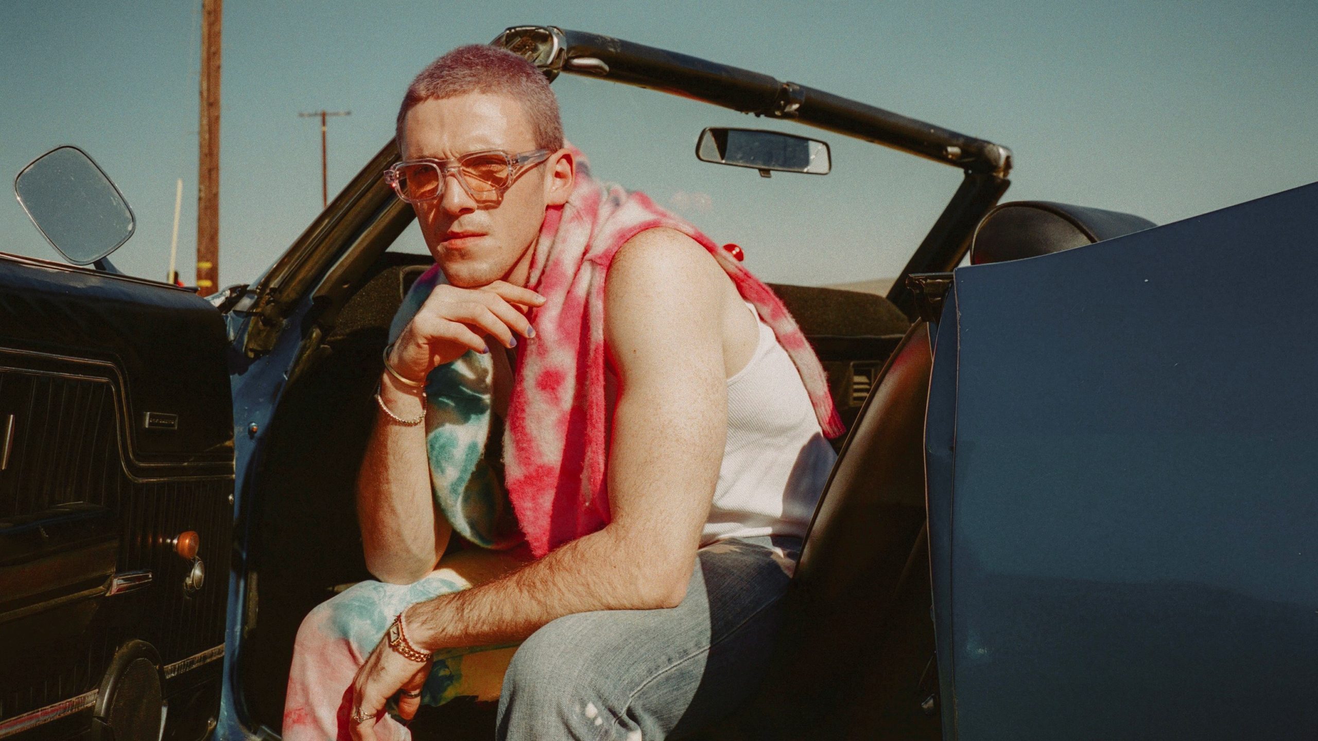 Man sits in convertible car with open door wearing a bright pink scarf and sunglasses