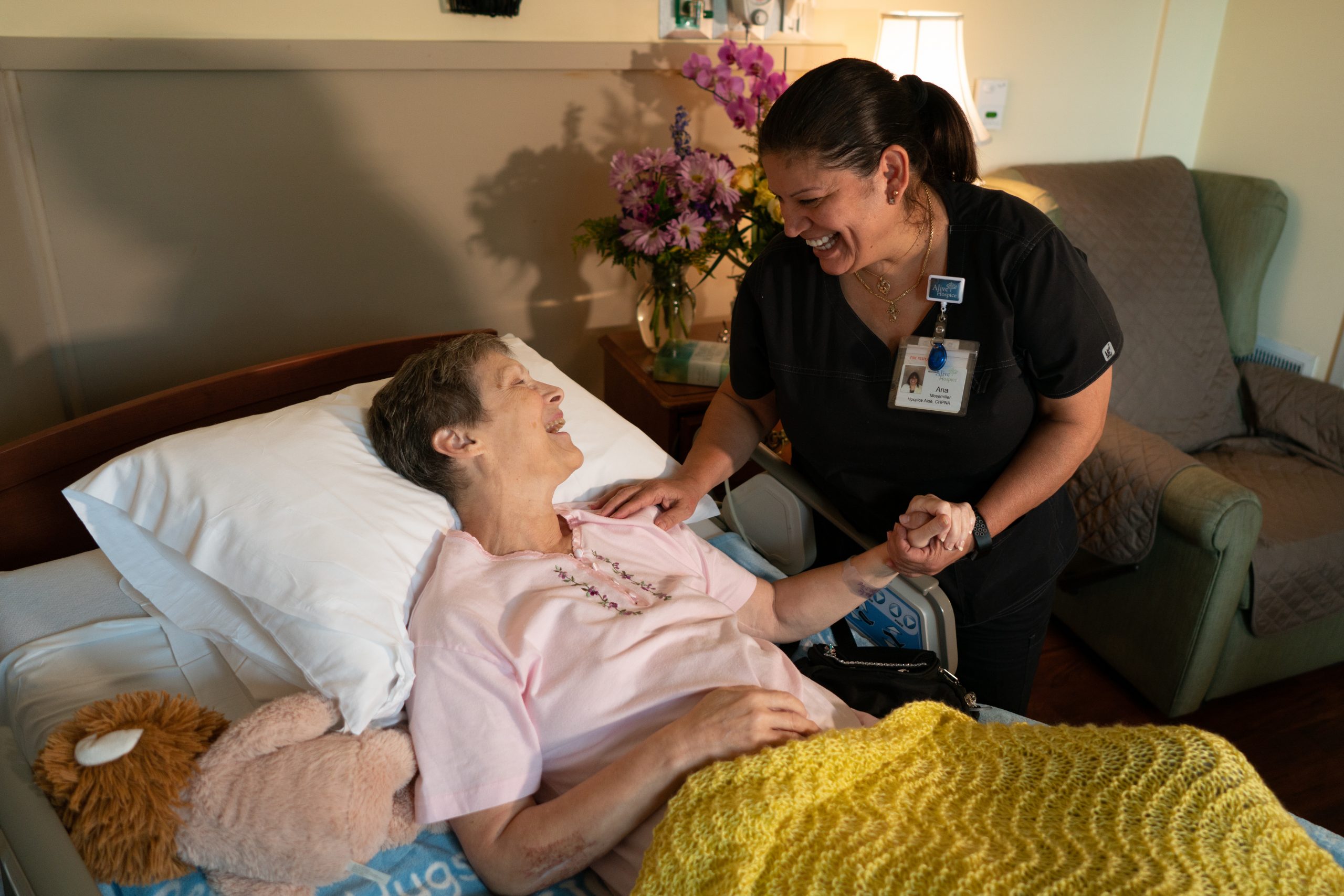 Photo of a smiling hospice nurse holding hands with and leaning over a smiling elderly female patient in bed.