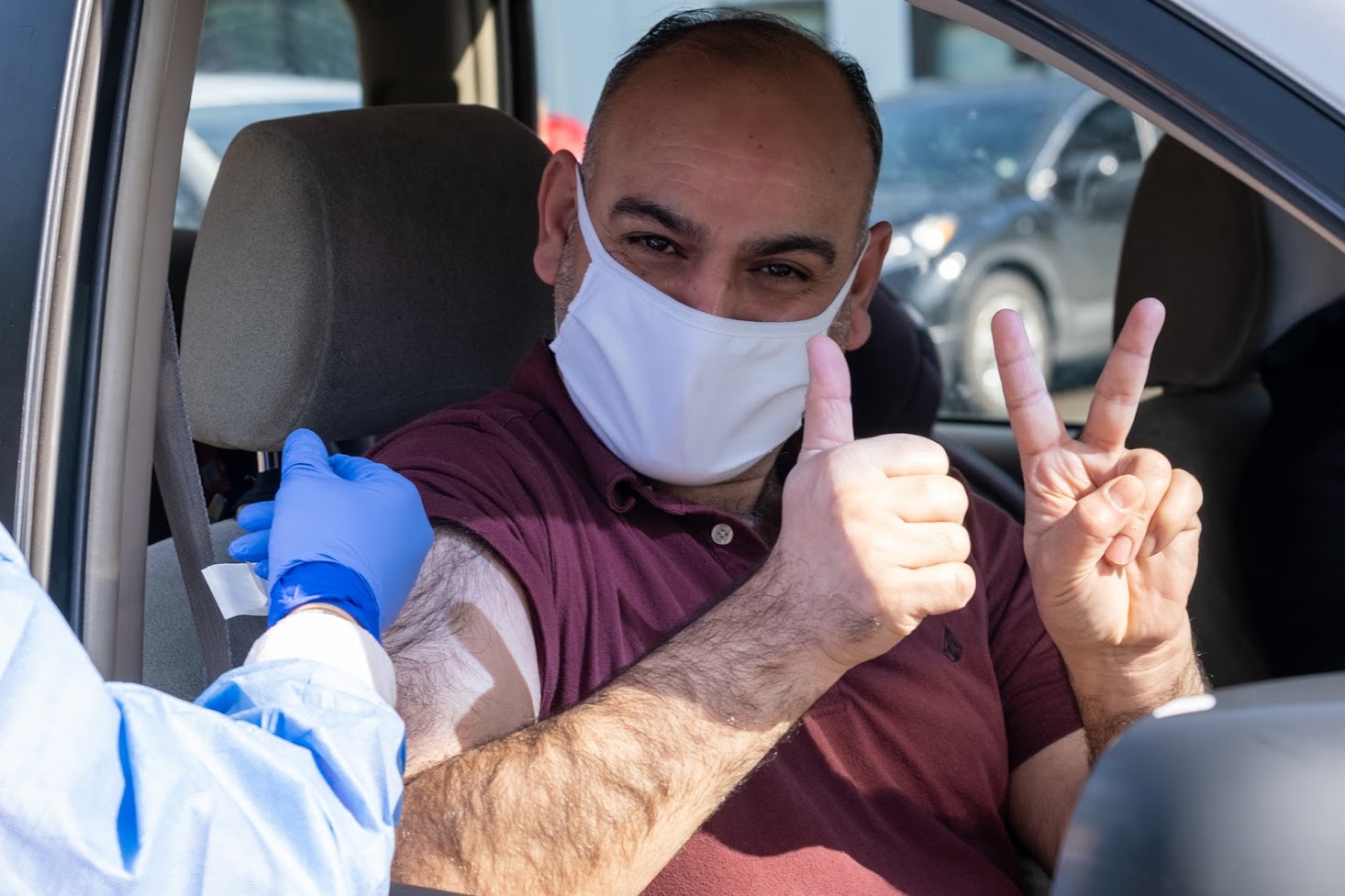 A vaccination patient in his car and wearing a white mask raises both thumbs after getting vaccinated by a medical worker