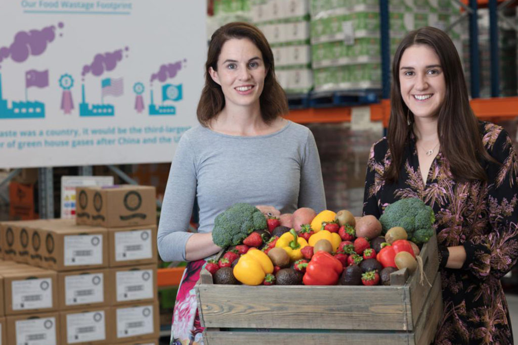 Two women standing behind a wooden box filled with fresh vegetables