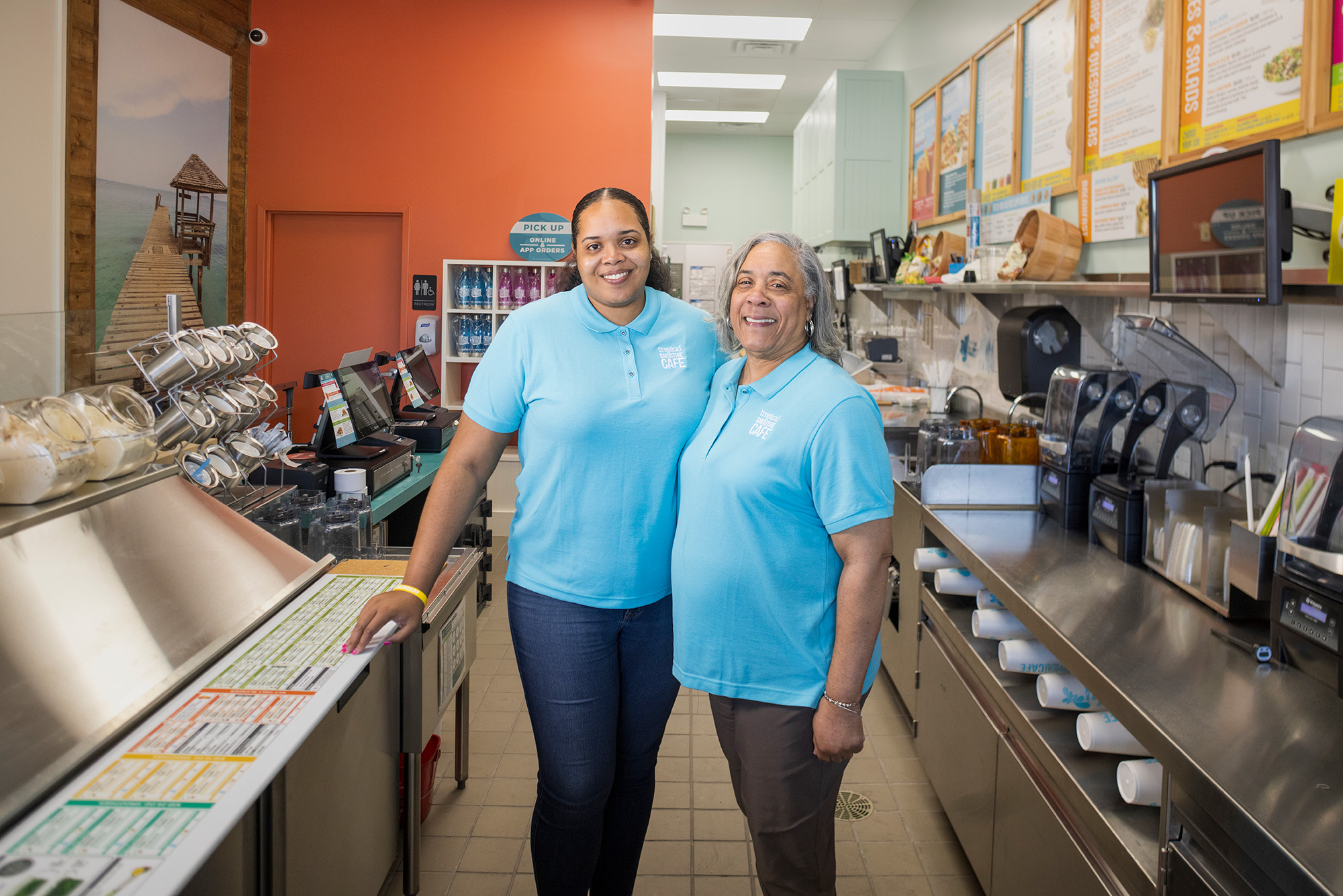 Two women in matching blue shirts stand together in a restaurant kitchen