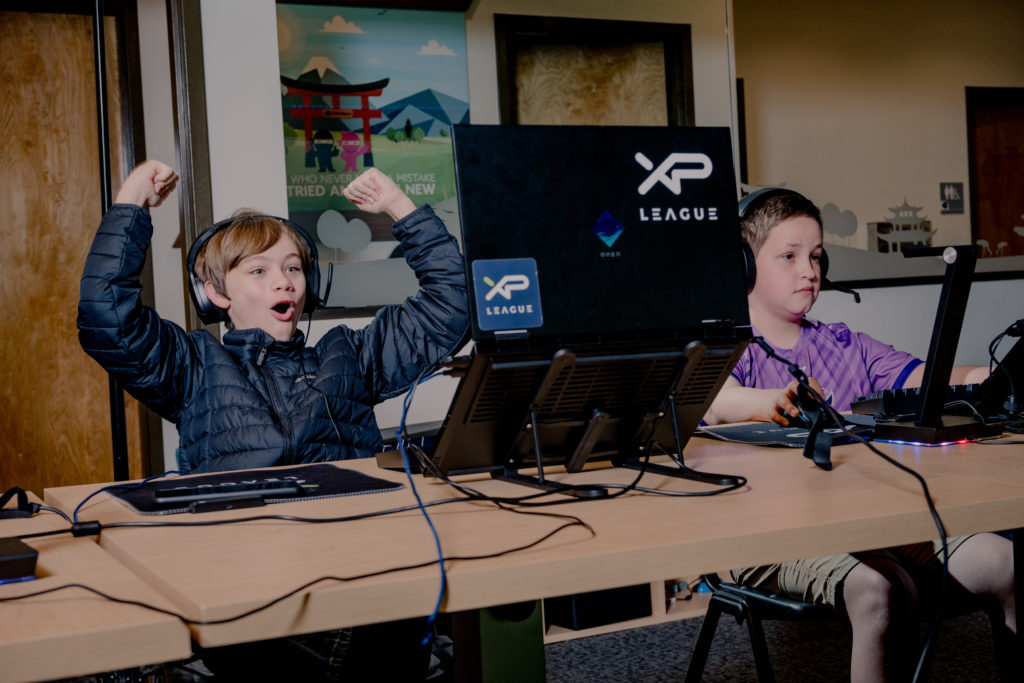 A boy wearing a winter jacket and headphones raises his arms in joy while watching his computer screen during an esports game.