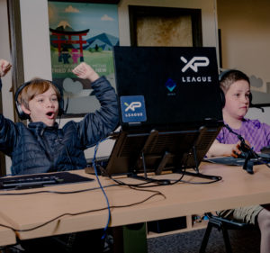 A boy wearing a winter jacket and headphones raises his arms in joy while watching his computer screen during an esports game.