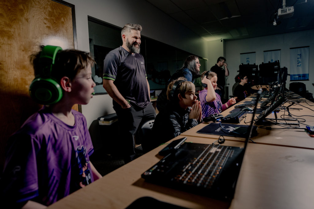 Jay Melamed and the mother of an XP League player stand behind a team of five teenage boys who are sitting in front of laptop screens and playing Fortnite. 