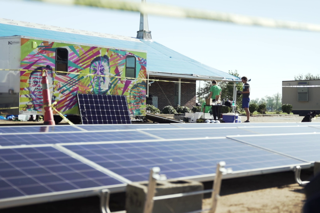 Image of solar panels laid out on the ground, with a colorful solar-powered trailer in the background. 
