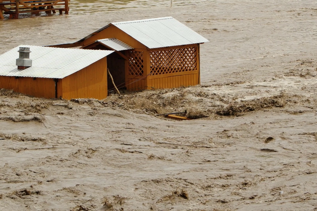 Image of muddy water flowing by two partially submerged buildings. 