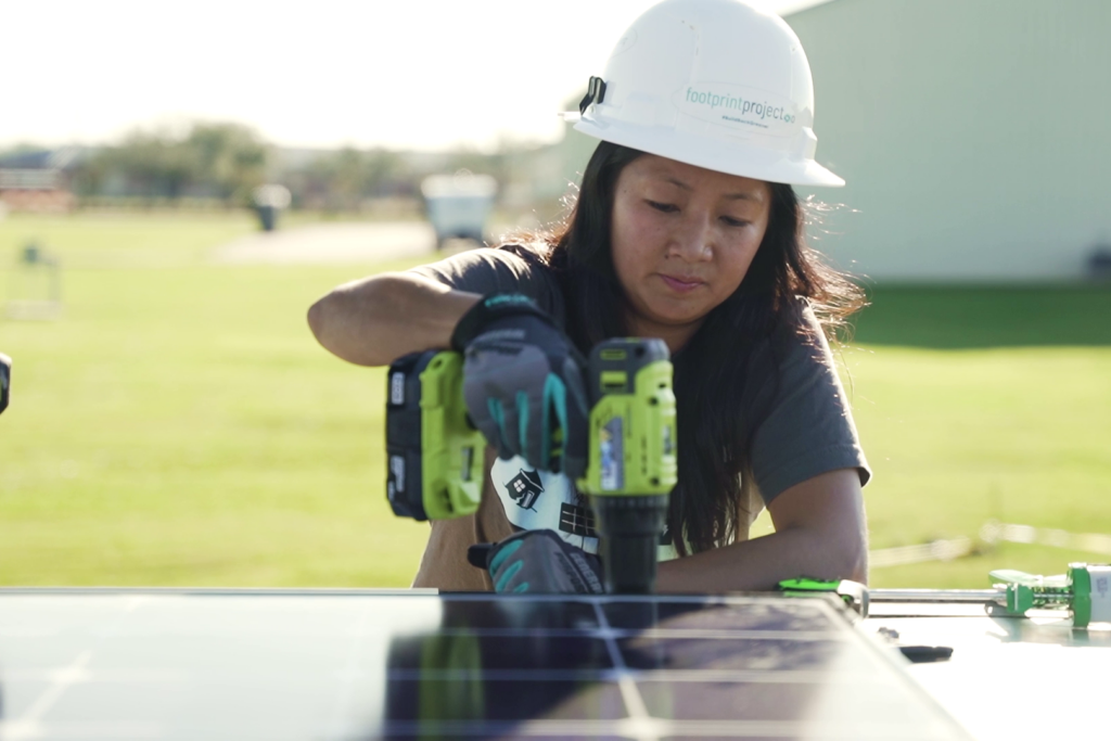 Image of a young woman wearing a hardhat and working on the top of a solar-powered trailer with a handheld power tool.  