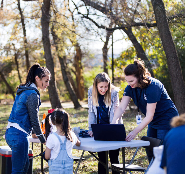 Woman and three girls around a laptop computer on a picnic table in a park