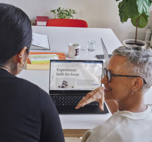 Viewed from behind, two people talk with each other in front of a laptop computer in an office