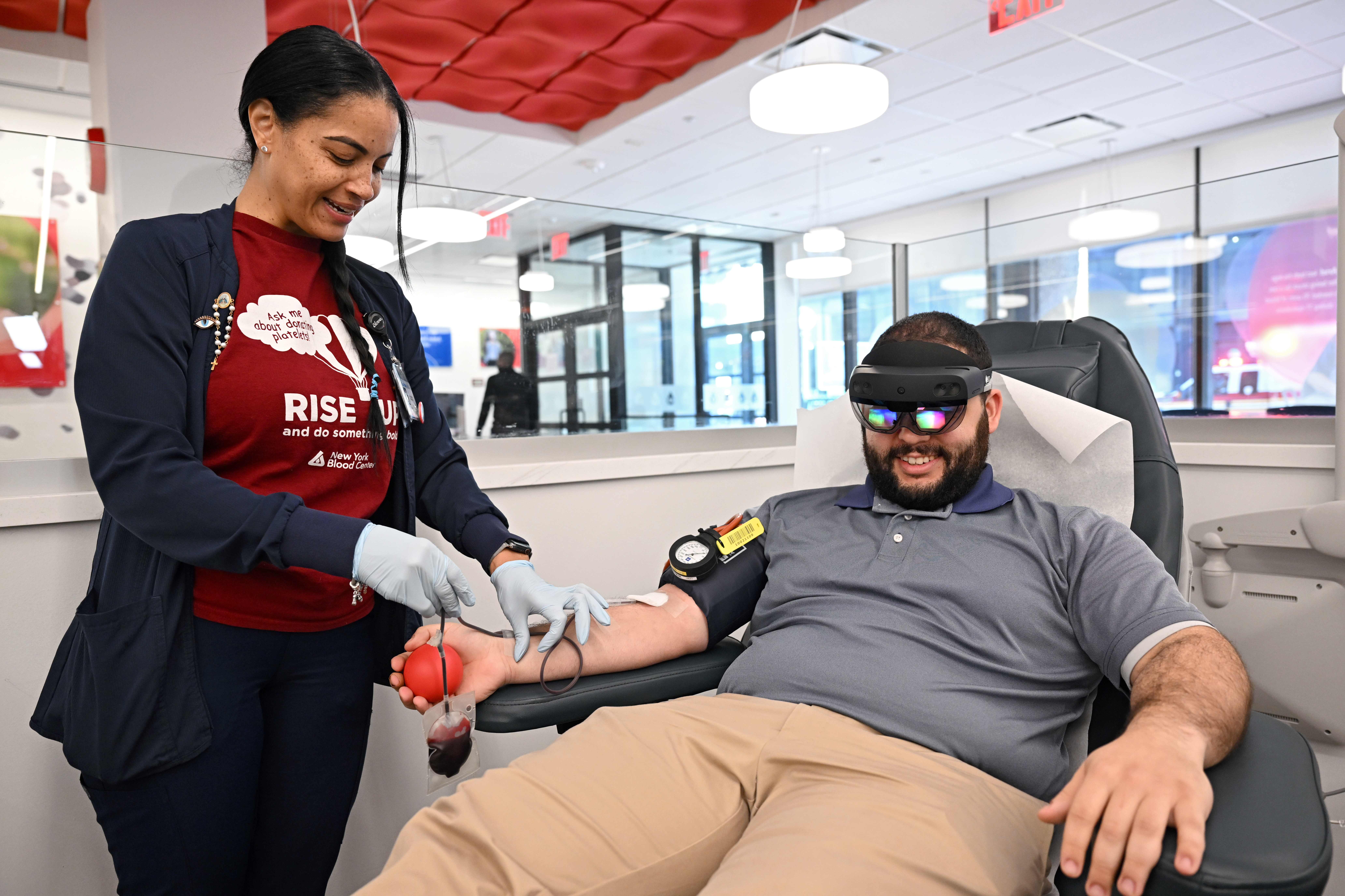 A staff member at a blood donation site collects blood from a seated donor who is wearing a HoloLens 2 headset. 