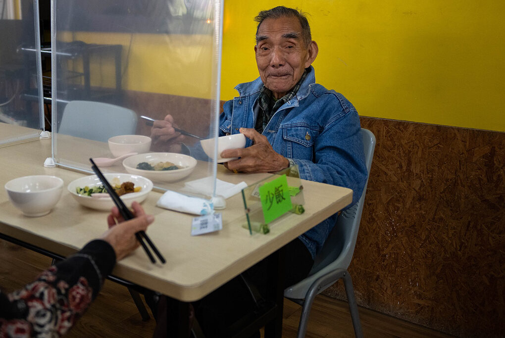 Two elderly men sit across a table, each holding a bowl and chopsticks 