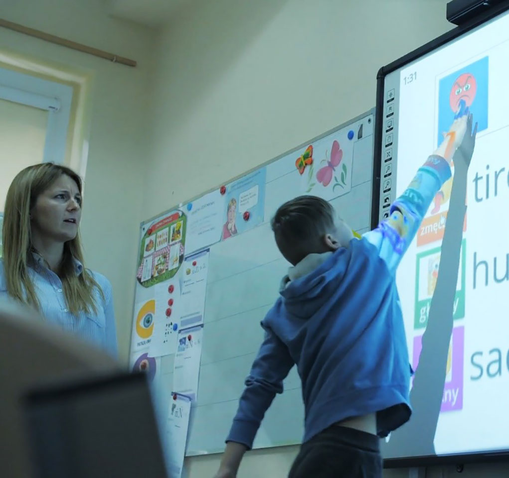 Teacher watching as student reaches up to tap one of the emotions displayed on a whiteboard