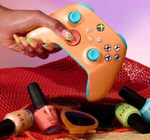Woman's hand with long painted fingernails holds an Xbox controller above four bottles of fingernail polish and a pair of sunglasses