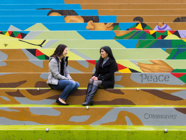 Two women sit outside on stairs while talking