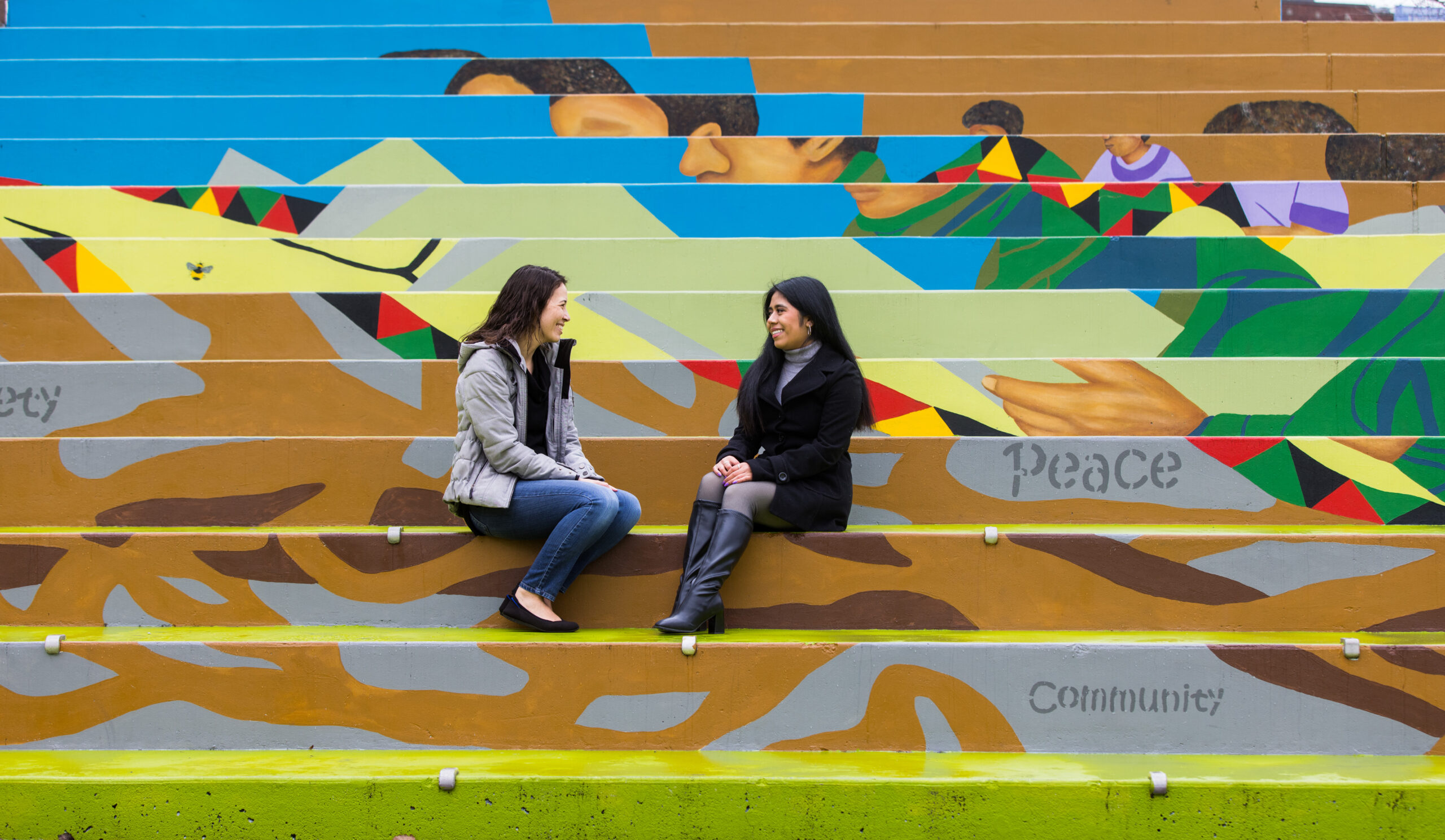 Two women sit outside on stairs while talking