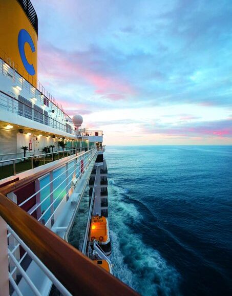 A cruise ship underway, shown from the starboard side, sailing toward an ocean horizon at sunset. 
