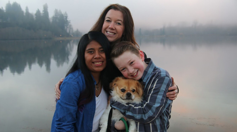 A mom and her two kids stand smiling by a lake with their dog