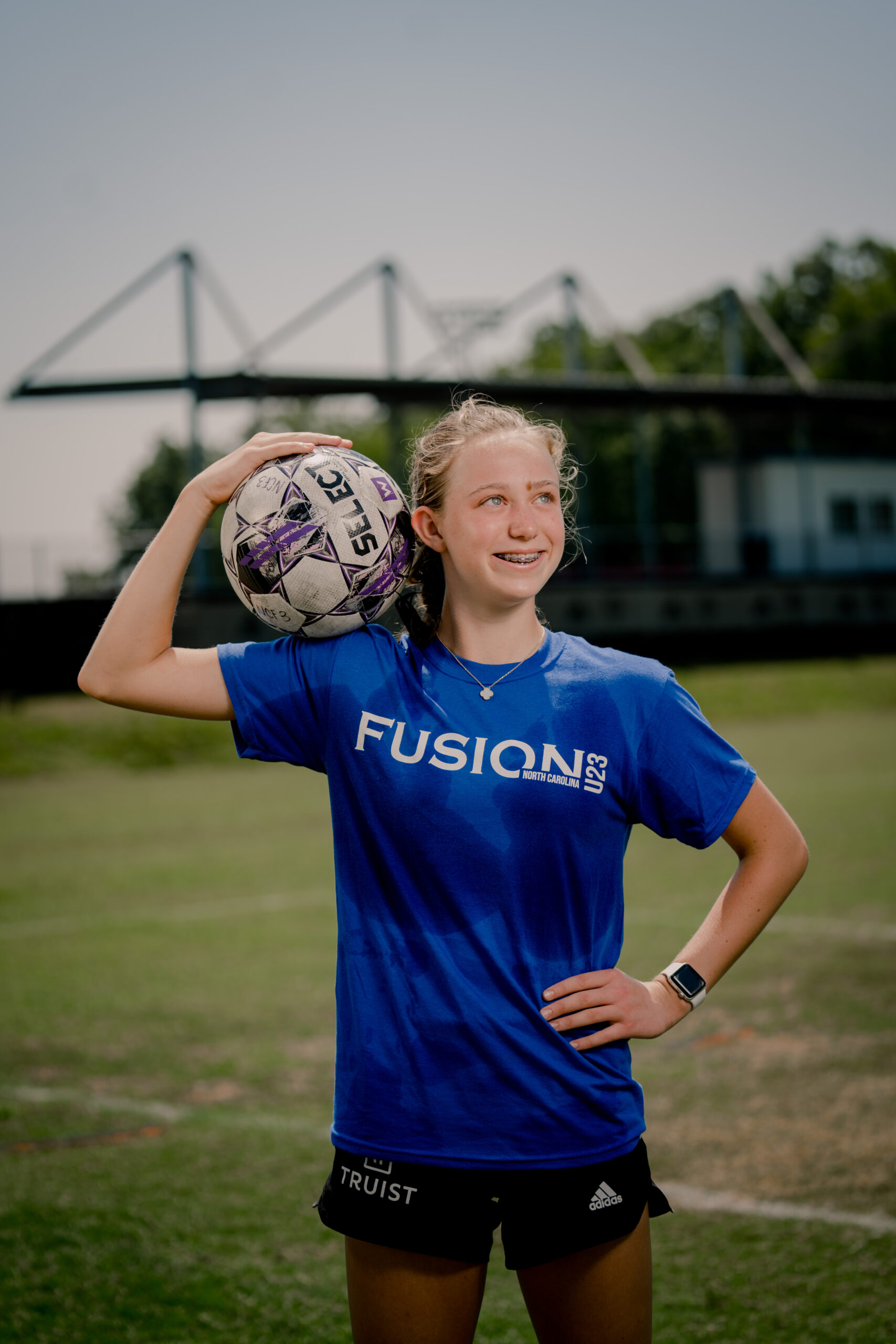 Sophie Wyshner stands on a soccer field and holds a soccer ball on her right shoulder. She