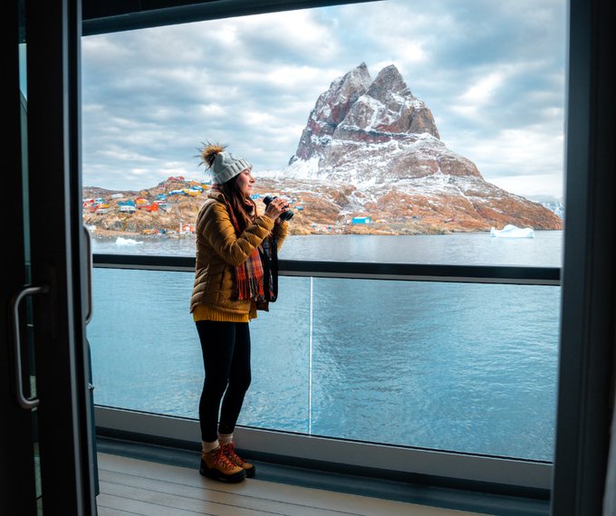 A cruise ship passenger holding binoculars stands on deck and looks out at water and a snowy island.