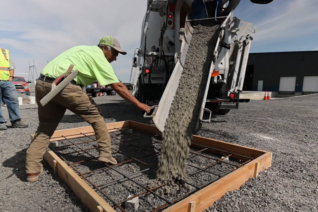 Construction worker pours concrete into a table-top size wooden frame.