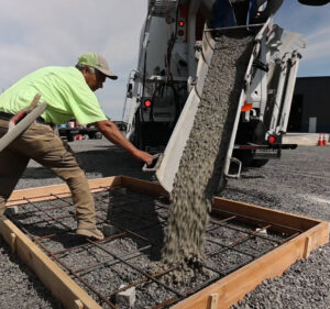 Construction worker pours concrete into a table-top size wooden frame.