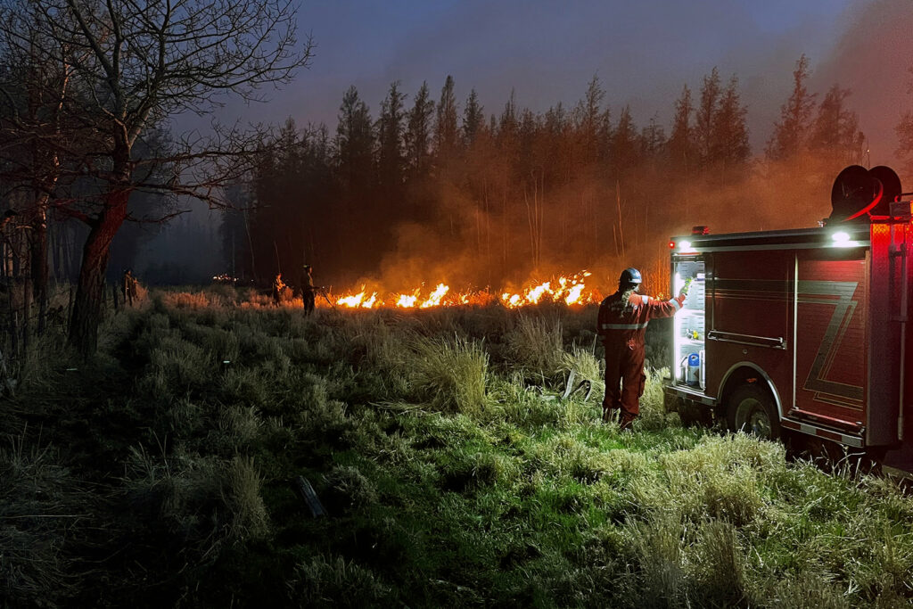 A firefighter stands at the back of an illuminated firetruck at night, with a fire off in the distance at the edge of a field