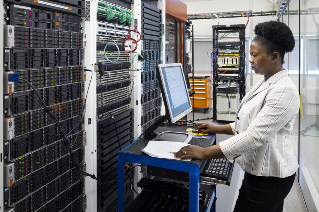 A woman works on a mobile computer station next to a server rack