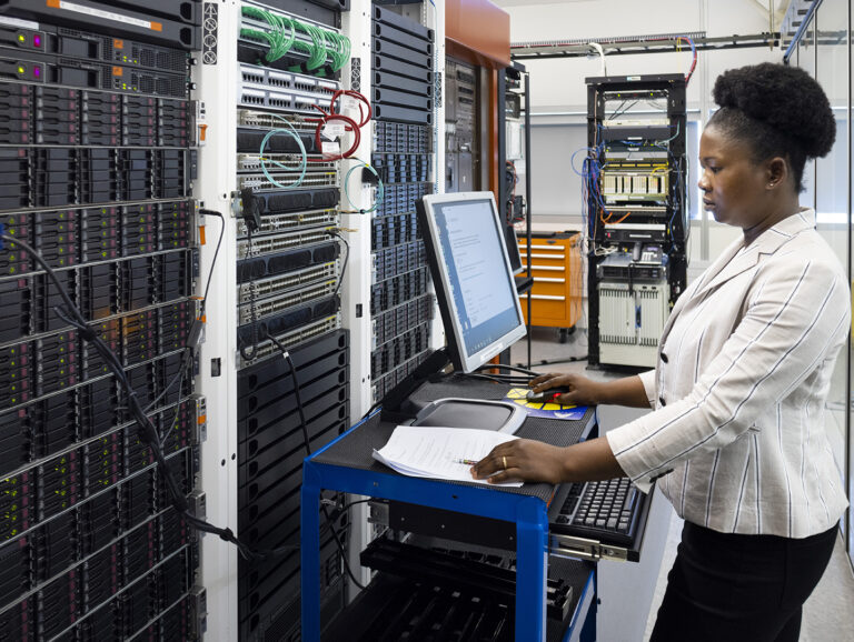 A woman works on a mobile computer station next to a server rack