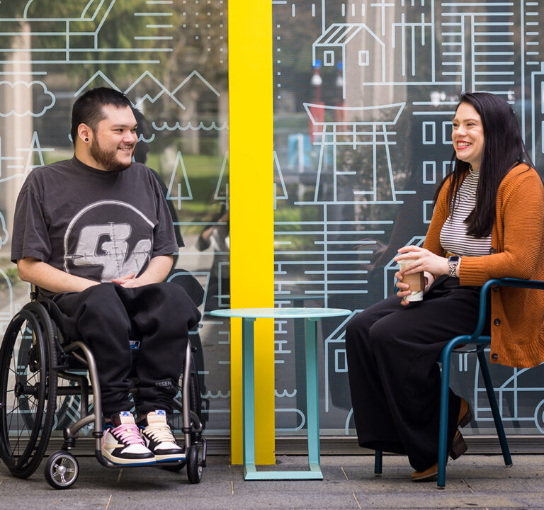 Man in a wheelchair conversing with a seated woman in an office lobby where walls are decorated with urban and rural scenes