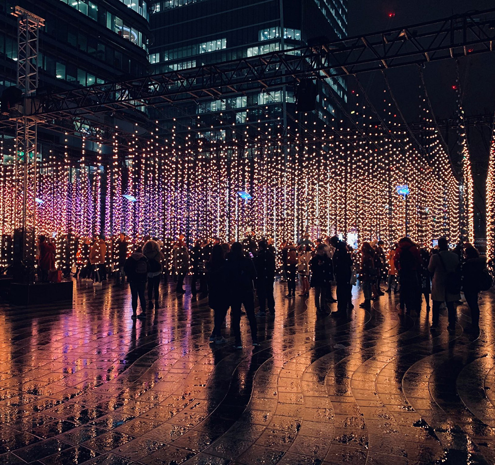 A crowd of people in a city square at night, illuminated by vertical strings of lights