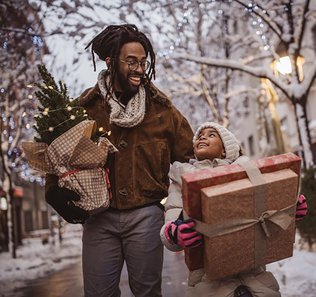 Father and child carrying gifts and a Christmas tree as they walk down a city street dusted with snow