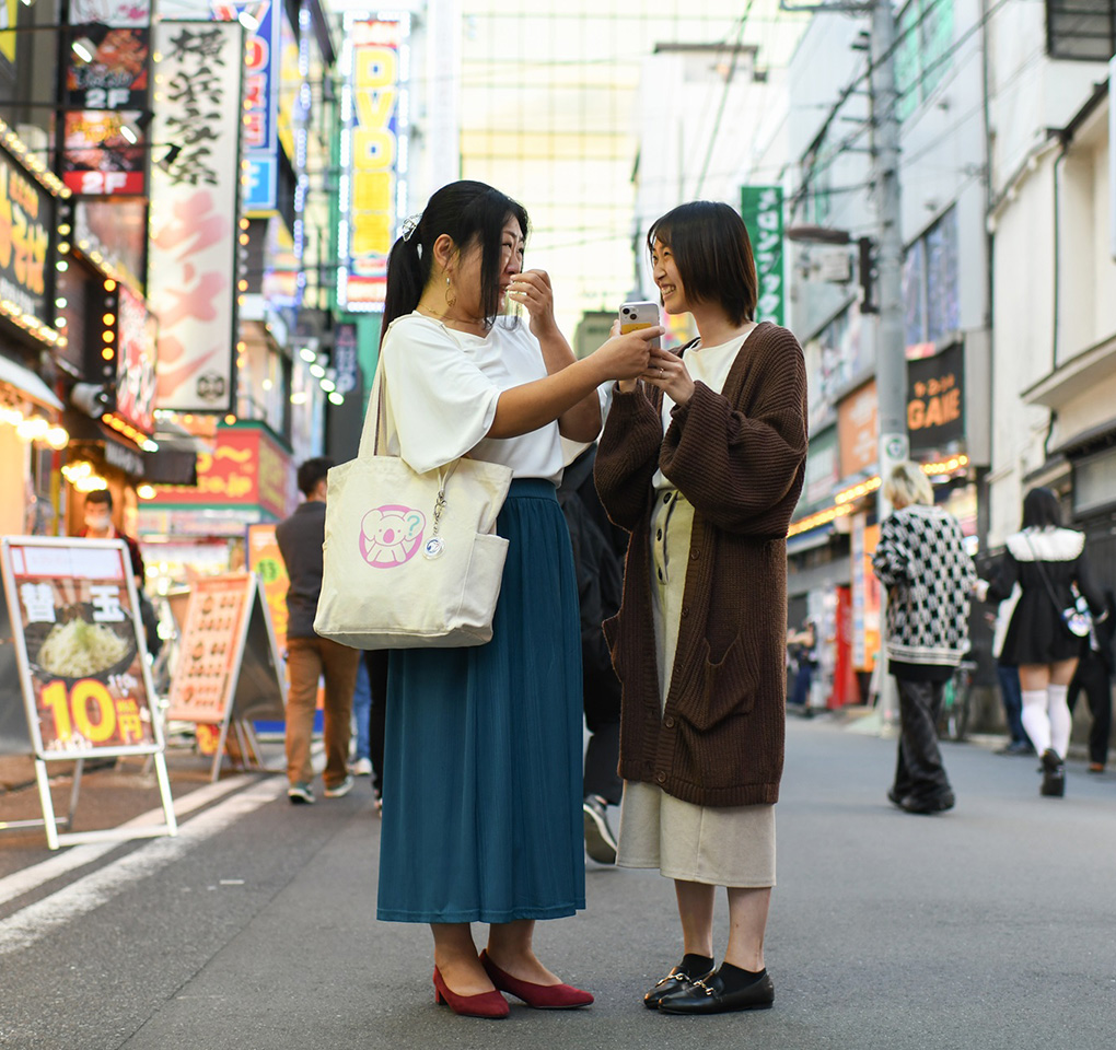 Two Japanese women stand in a market sharing what's on on of their phones