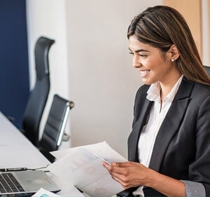 Woman smiling in an office as she goes through papers and interacts with a laptop computer