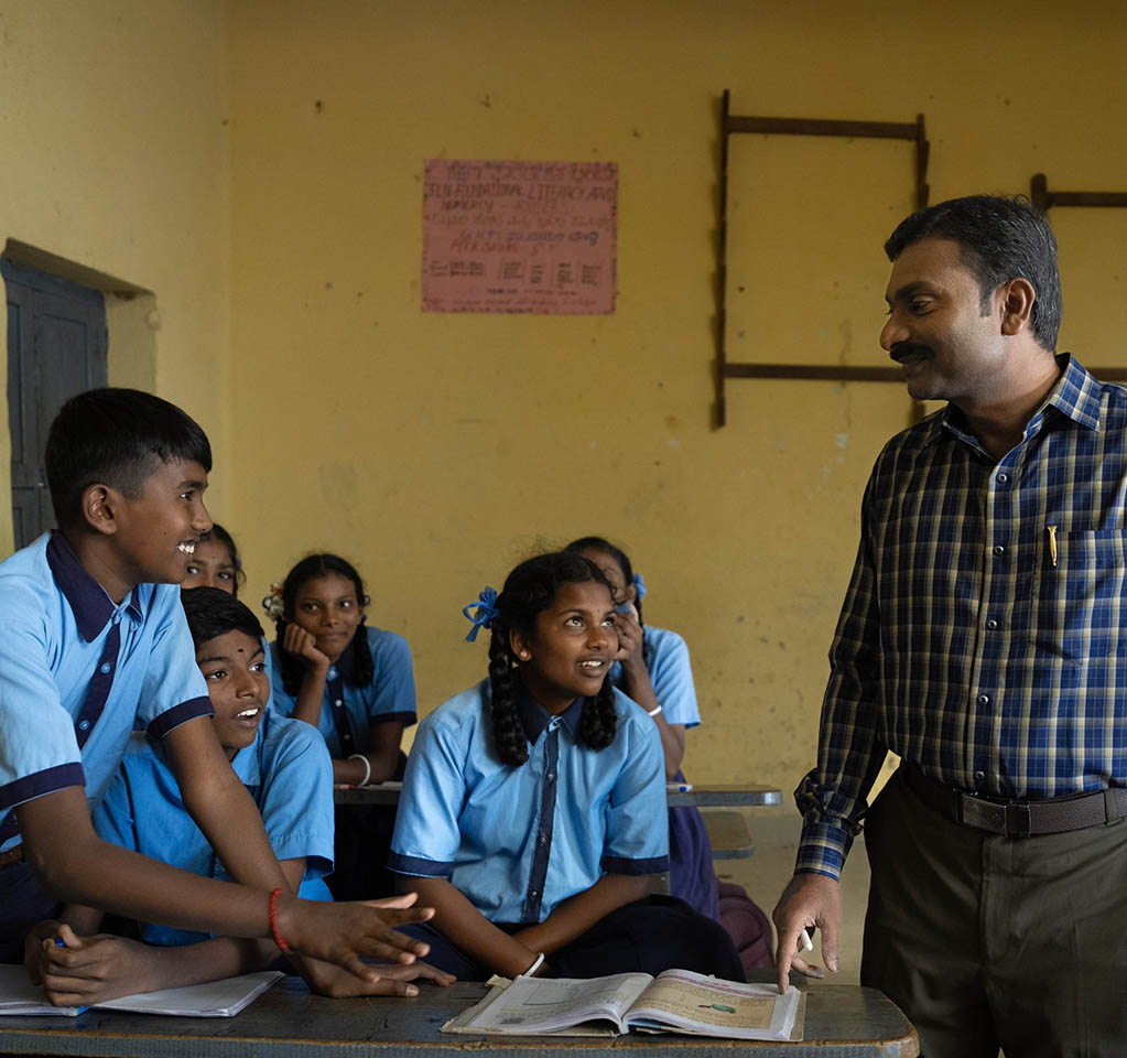 A male teacher in a blue plaid shirt interacting with students in blue uniforms in a classroom  
