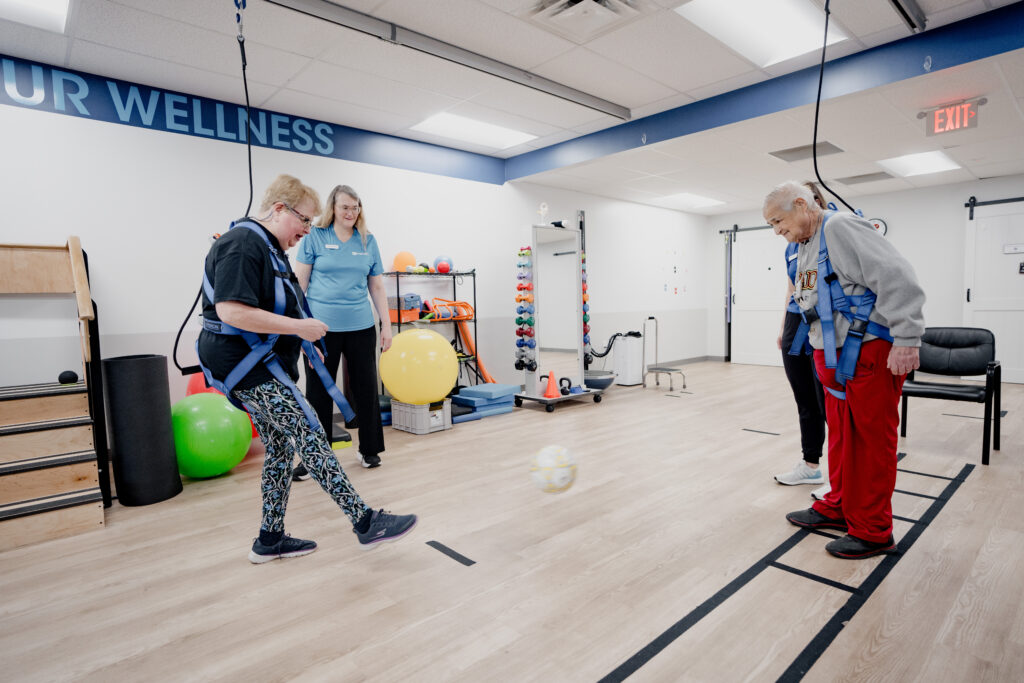 A man and a woman in safety harnesses kick a soccer ball together in a physical therapy clinic.