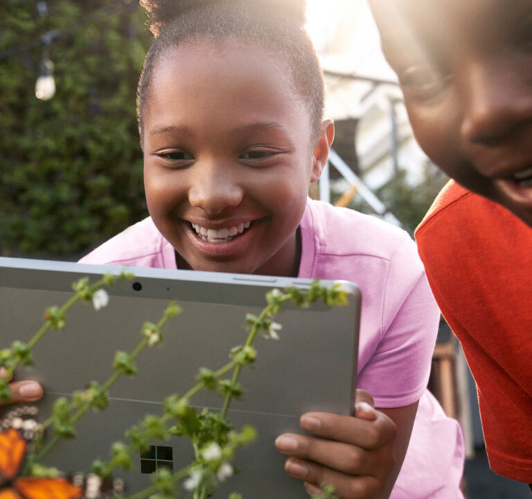 Two students smiling as one of them takes a photo of a plant with her tablet device