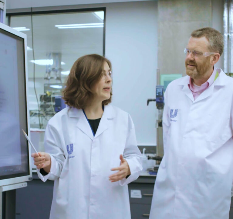 Woman explains something to a man, both wearing lab coats, while pointing to what's on a large screen