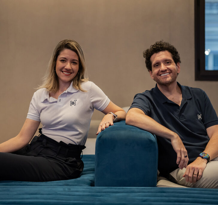 Christianne Bariquelli and Marcos Albino Rodrigues sitting on a sofa, facing the camera. Both are smiling and wearing polo shirts with the B3 logo.