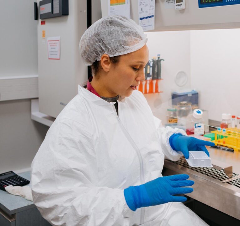 A woman in a lab coat, with her hair in a net and her hands in blue gloves, holds a sample in a lab.