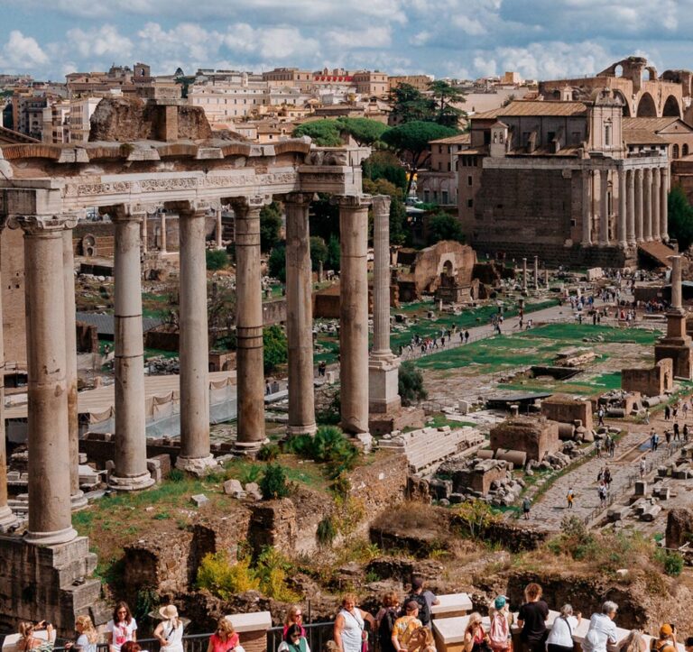 In the foreground, a crowd of tourists is looking over a scene of ancient ruins, with columns and various stone structures under a blue sky.