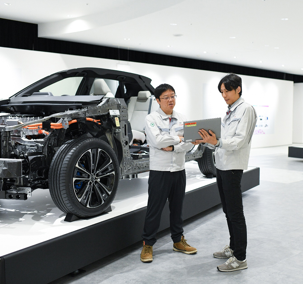 Two men look at a laptop next to a car display with the vehicle body peeled off to show the engine and other internal components.