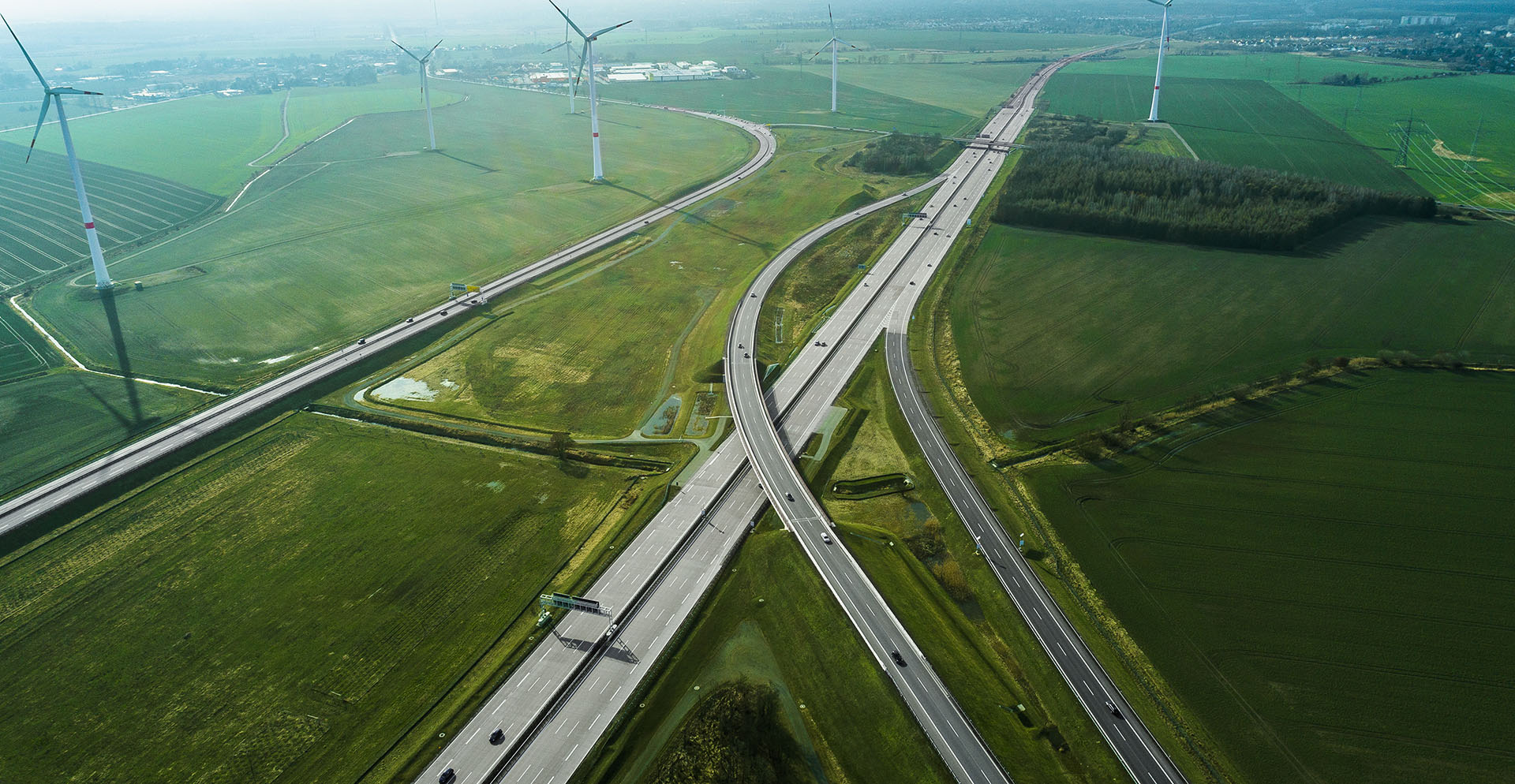 Aerial view of intersecting highways surrounded by vast green fields, with wind turbines scattered across the landscape and small buildings visible in the distance.