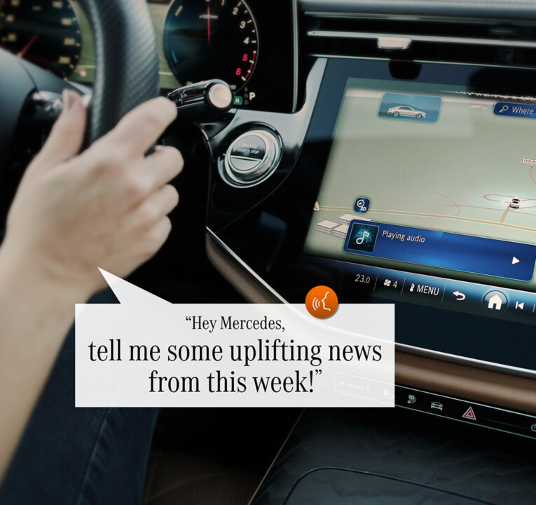 Dashboard of a Mercedes-Benz auto with a person's hand on the steering wheel and a caption from the driver saying Hey Mercedes, tell me some uplifting news from today.