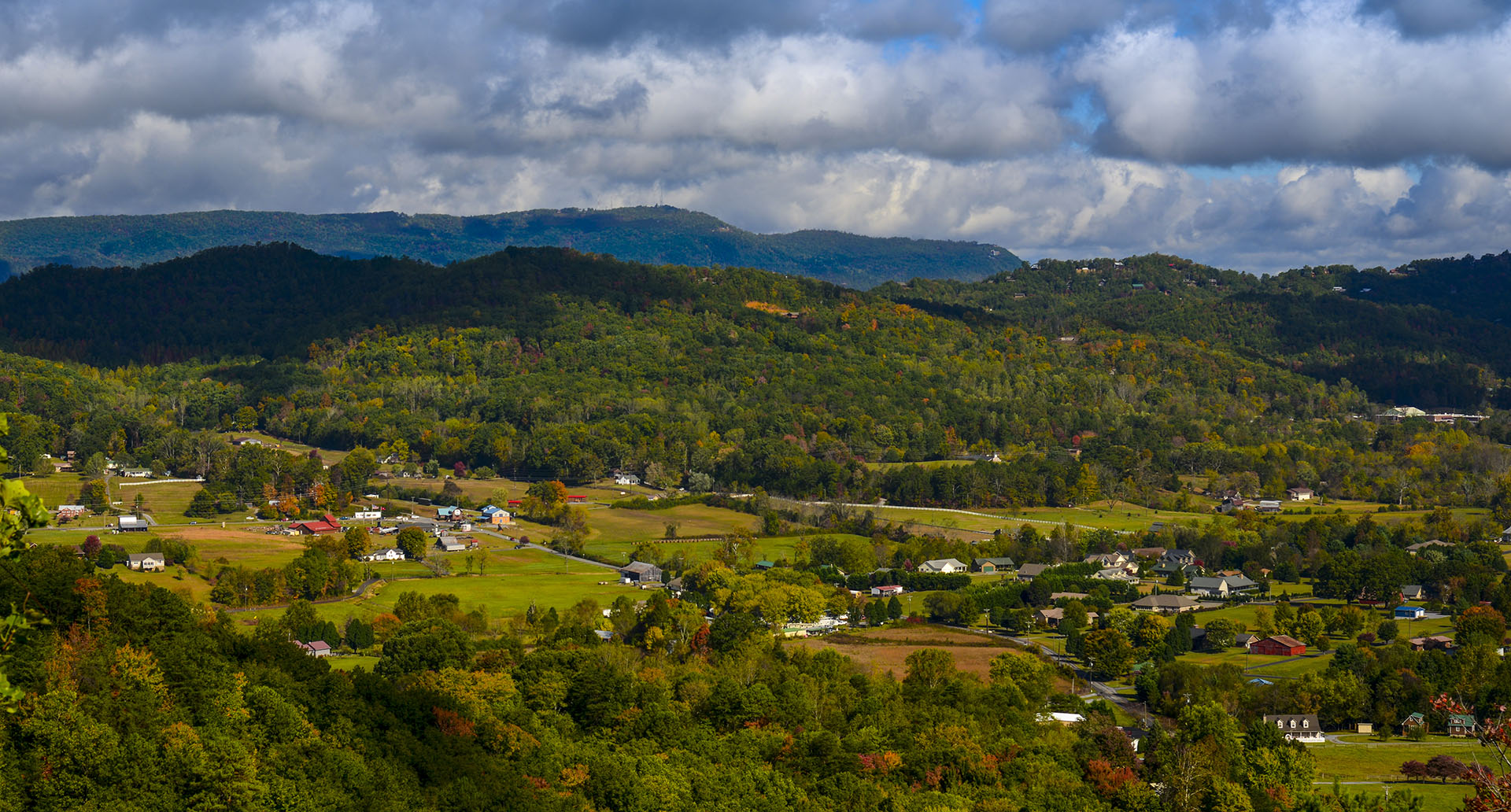 A lush green valley dotted with houses and farms, surrounded by forested hills and mountains under a partly cloudy sky.