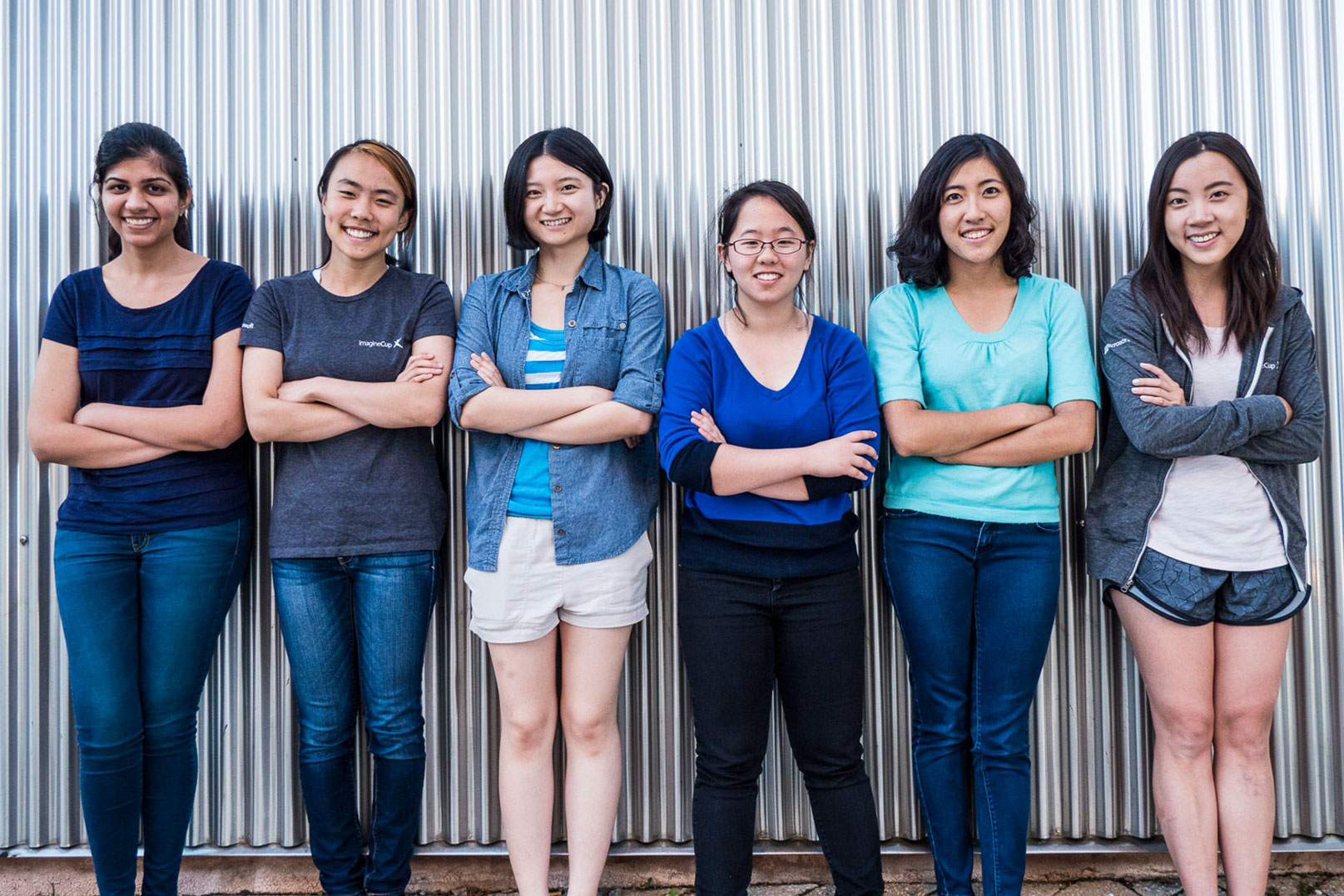 Portrait of the six students smiling and standing with their arms crossed against a corrugated metal wall