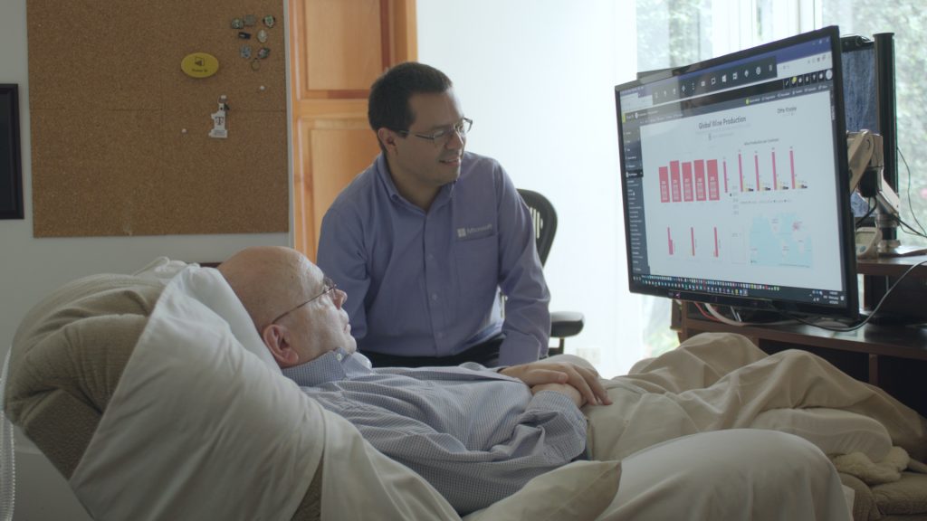 One man lies in a bed and another sits at his side, both looking at a computer screen
