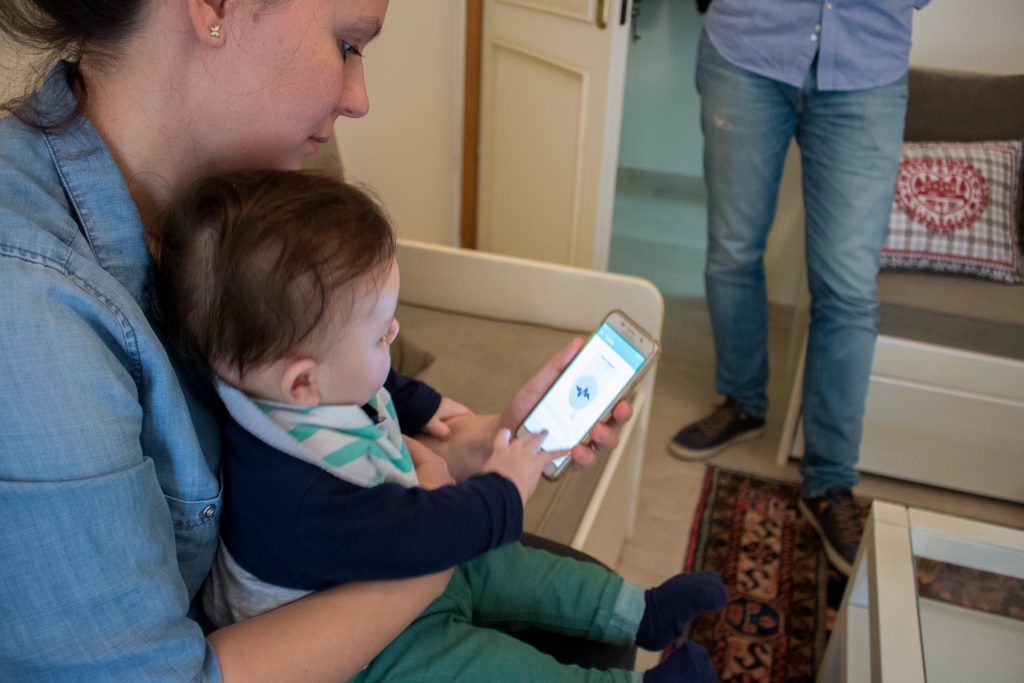 Мама держит ребенка и сомтрит в смартфон на котором открыто приложение для общения родителей и младенцев iCry2Talk (Греция)