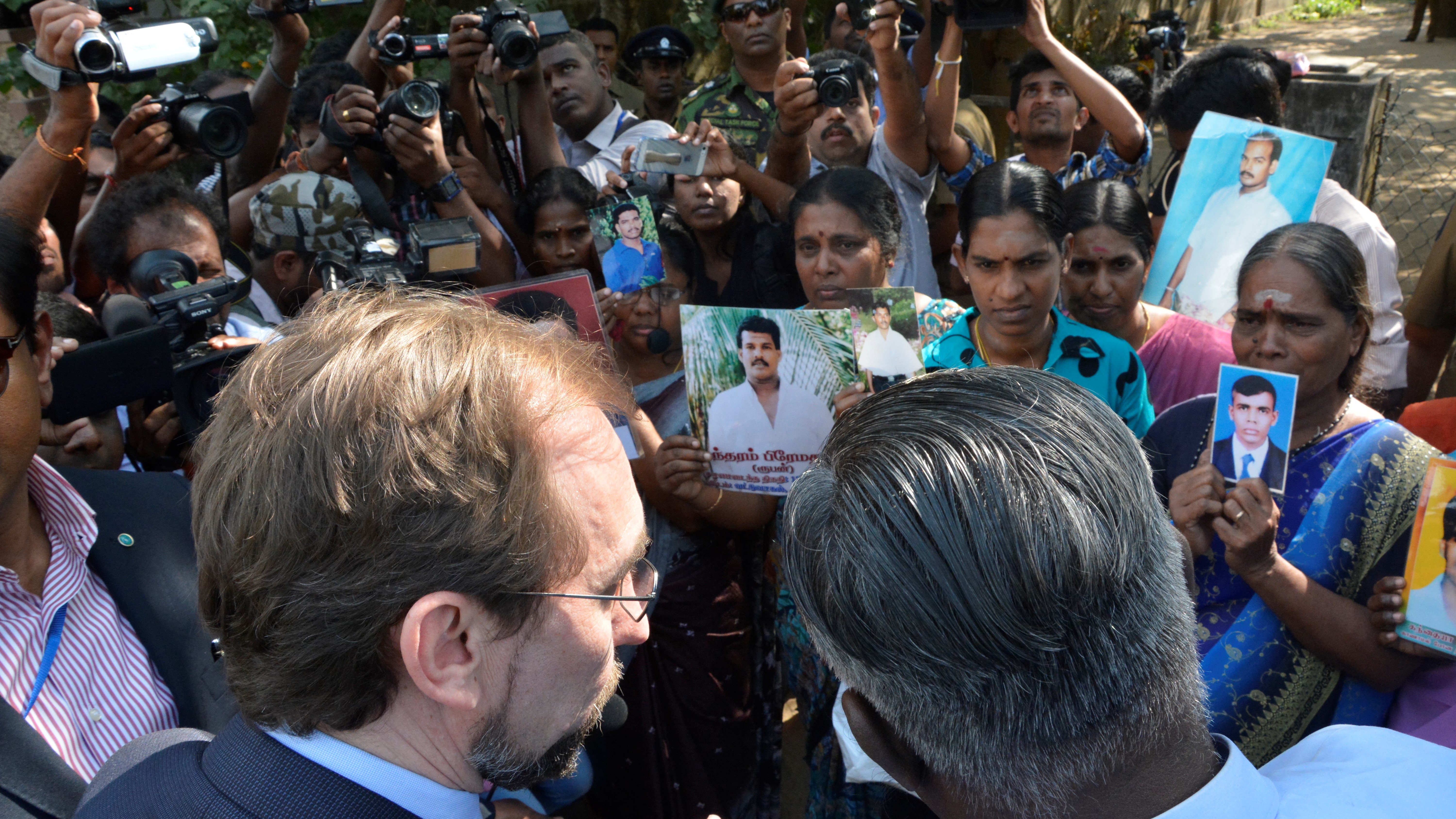 Photo of two men speaking to crowd of solemn people, some holding photos of loved ones