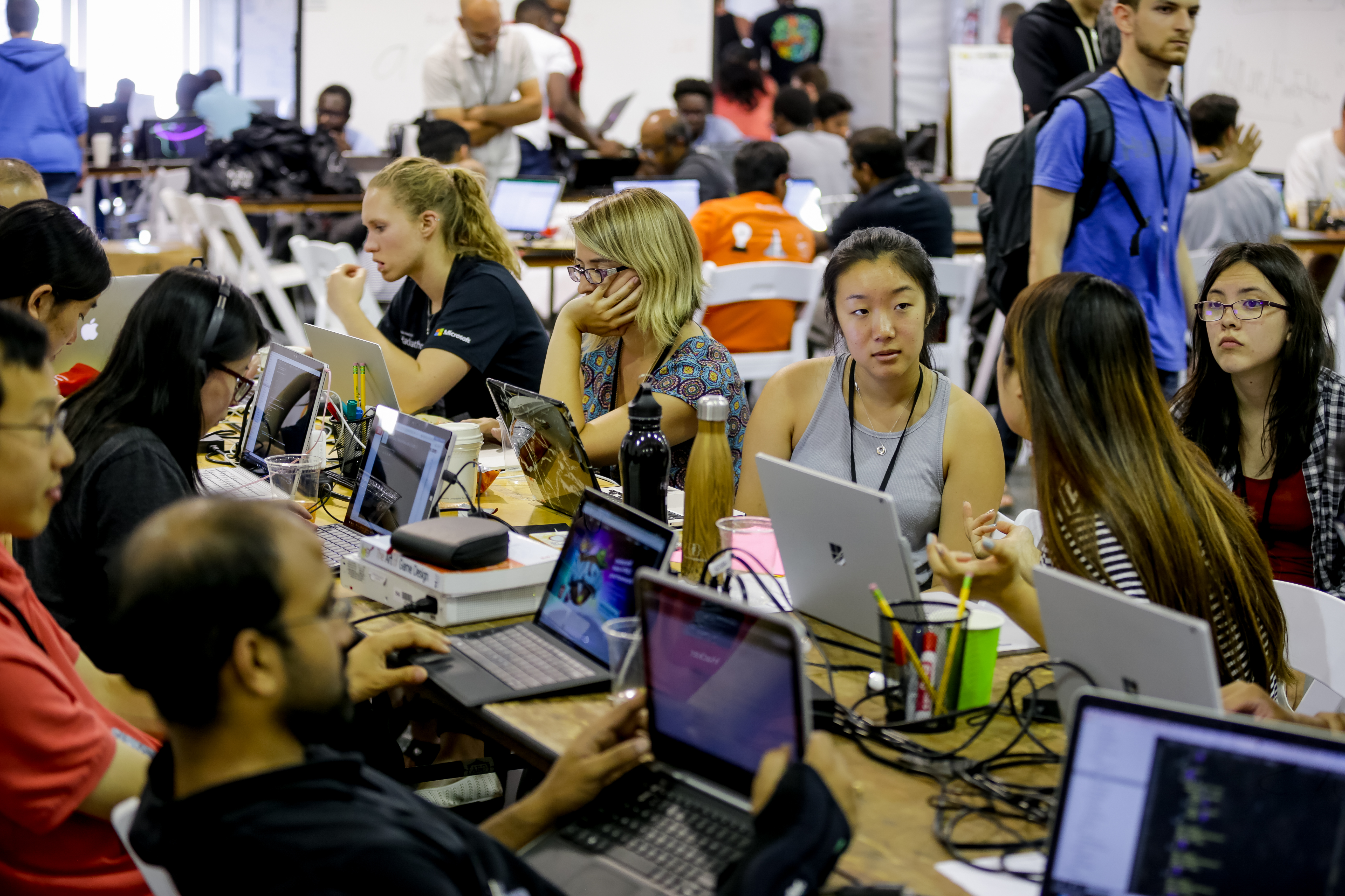 A group of Hackathon participants sit at long tables, staring at laptop screens or talking to each other in small groups.