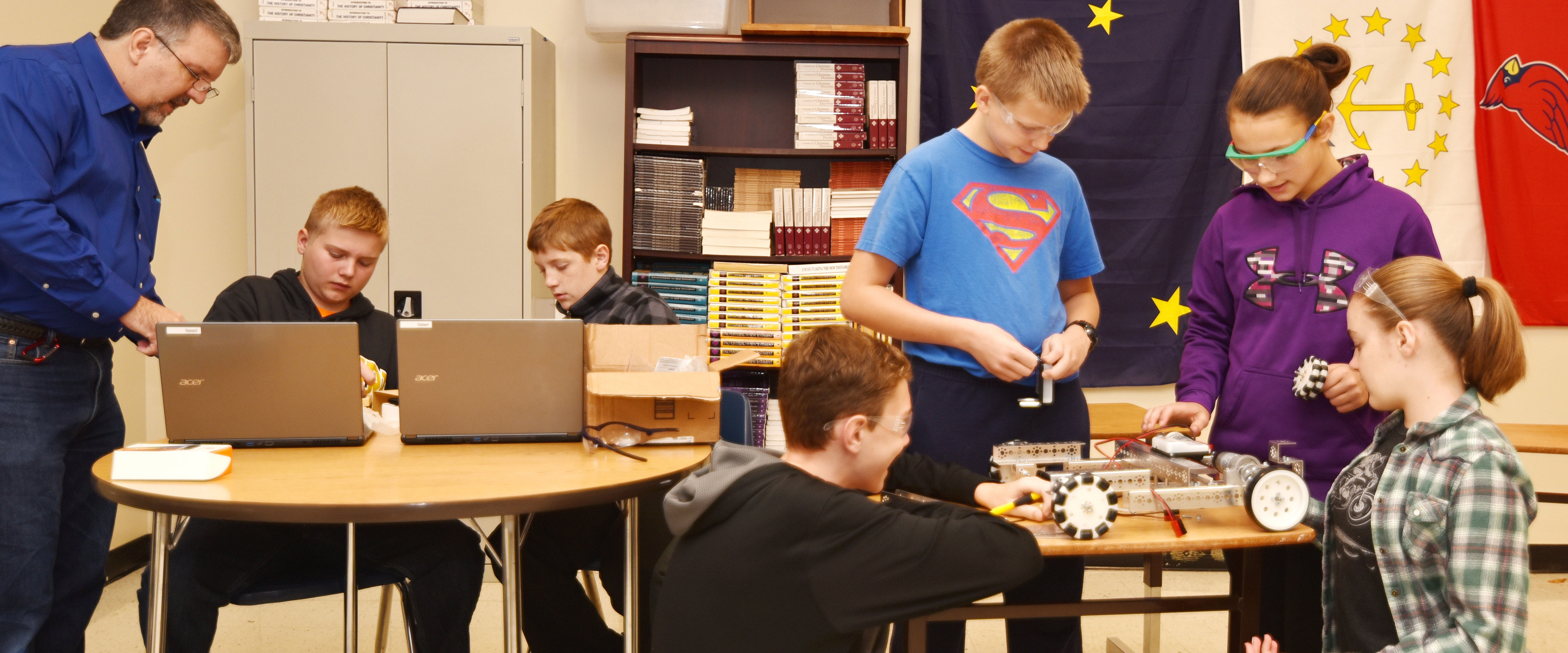 Photo of Kevin Sheck helping two boys who are working on laptops, while two more boys and two girls work on a robotics project at another table
